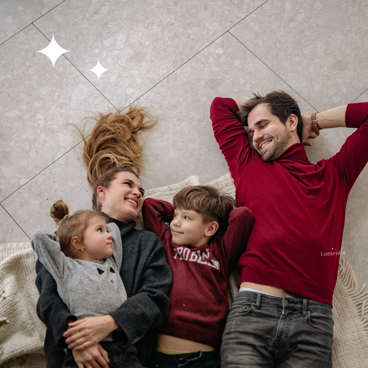 Family of four lying on tiled floor smiling at camera, wearing burgundy and gray clothing
