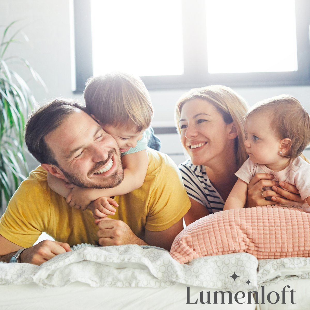 Happy family of four relaxing together on a bed in a bright, sunlit bedroom with plants in background
